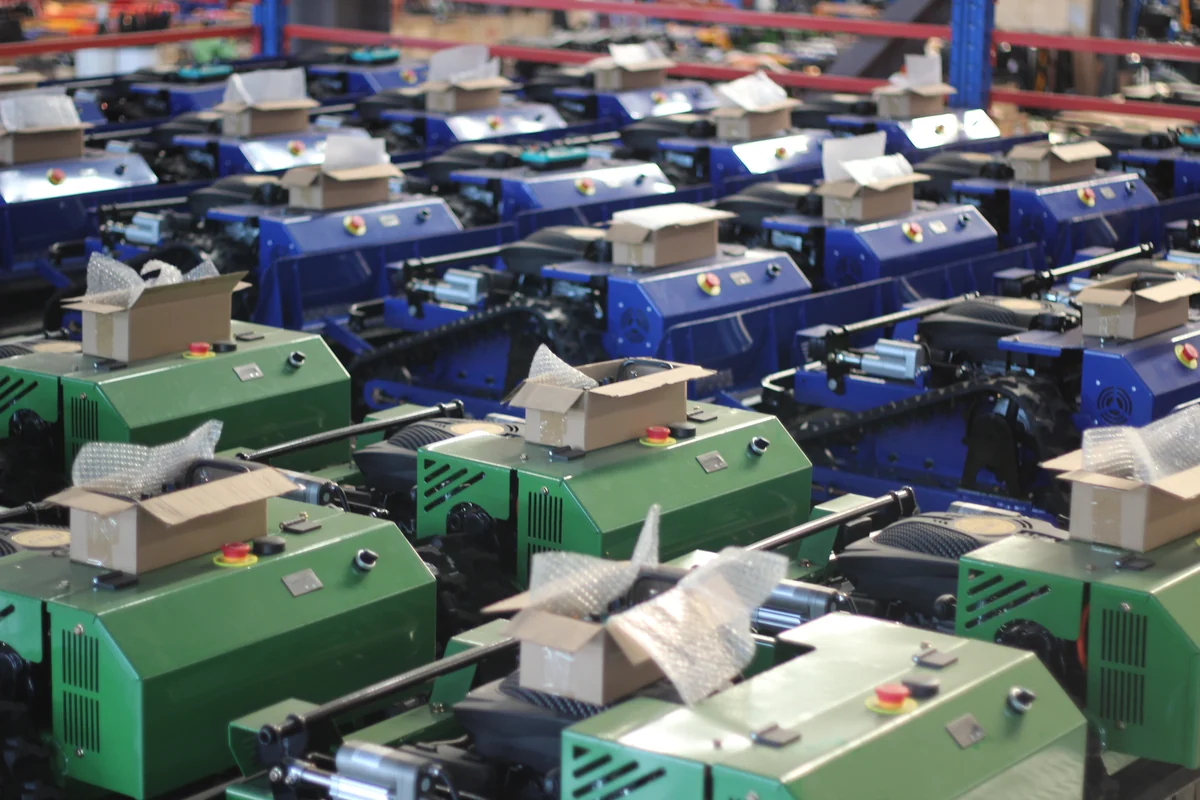 Rows of green and blue tracked mower chassis and control enclosures on the factory floor.